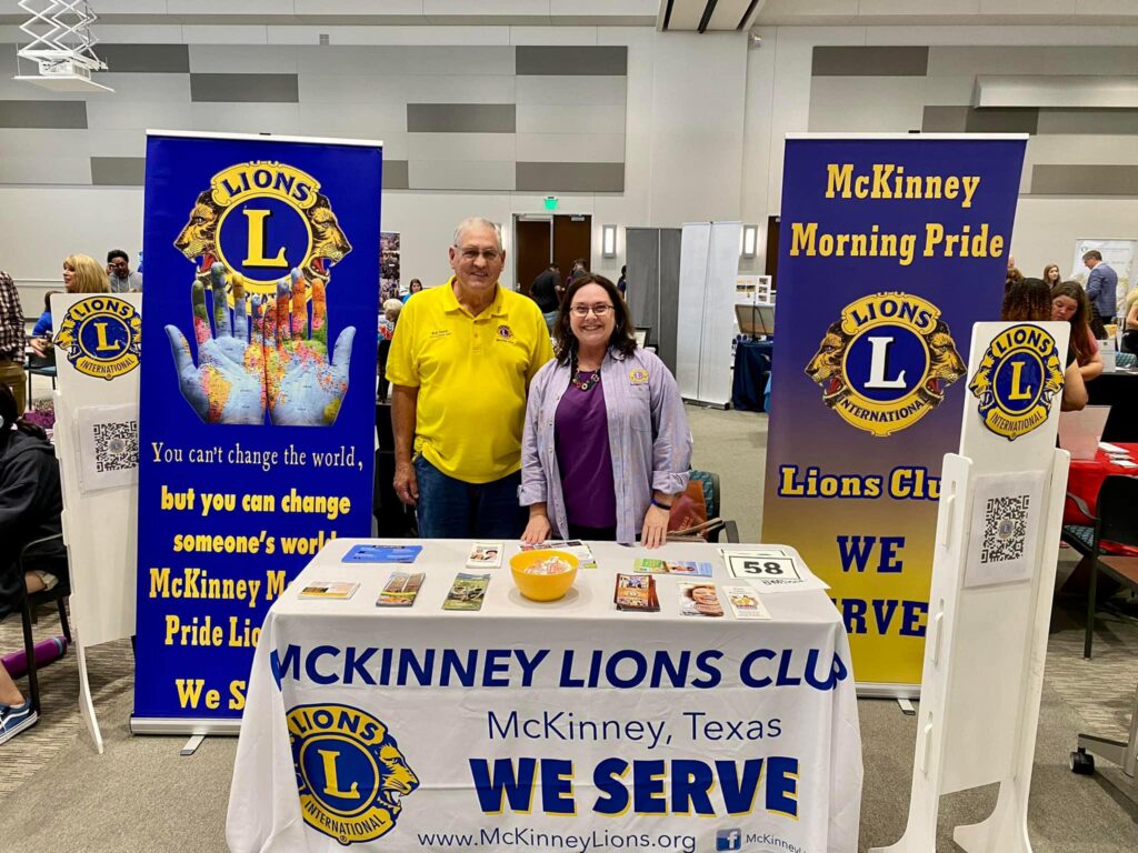 a color photo of a man and woman standing behind a table with information about the McKinney Lions Clubs with club banners behind them.