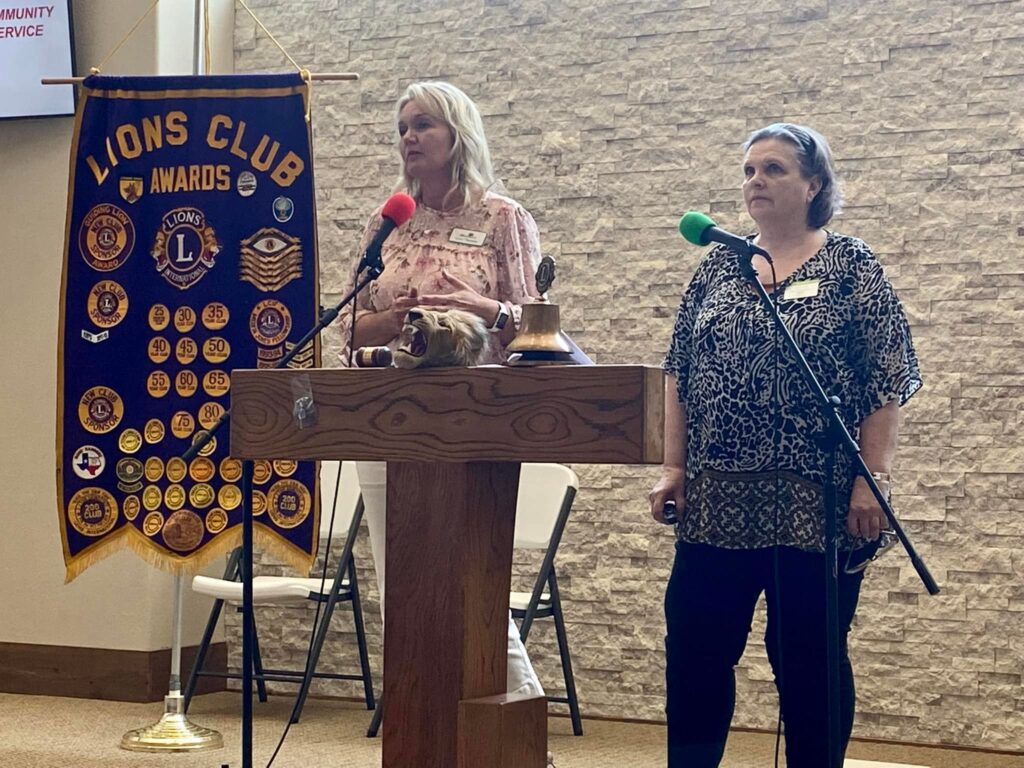 A color photo of two women at podium with a Lions Club banner next to them.
